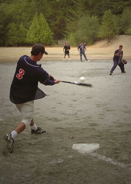 A boy in a black jacket hitting a baseball with a bat on a dirt field with several people and trees in the background.