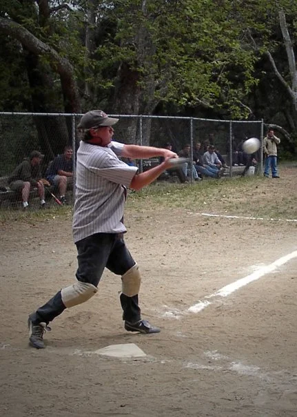 A young baseball player swinging a bat at a ball during a game at the field, with spectators watching from behind a chain-link fence.