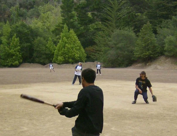 Young boy in black jacket swinging a baseball bat on a baseball field surrounded by trees and other children in the background.