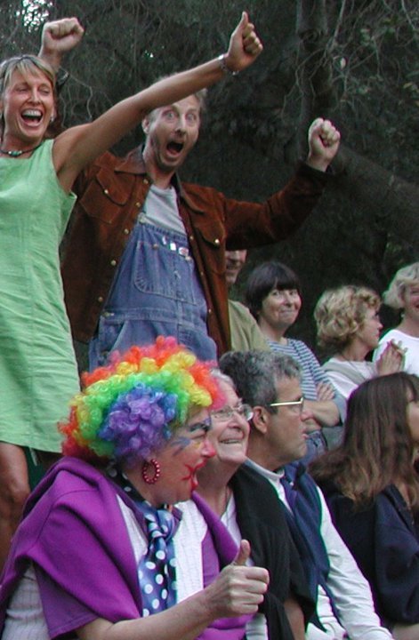 A group of people, including two with their arms raised and smiling, a clown with colorful hair, and others clapping or smiling at an outdoor event.