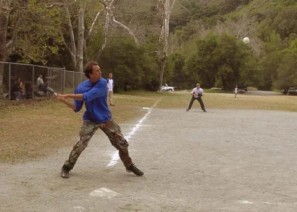 Person batting a baseball on a field with others watching and trees in the background.