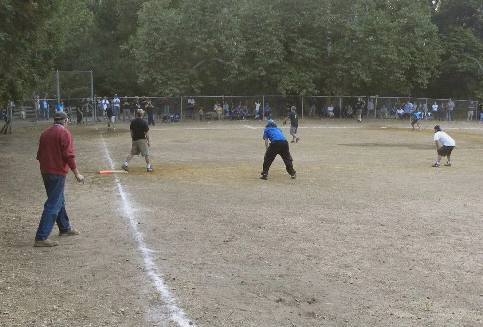 People playing baseball on a field with a chain-link fence and trees in the background.