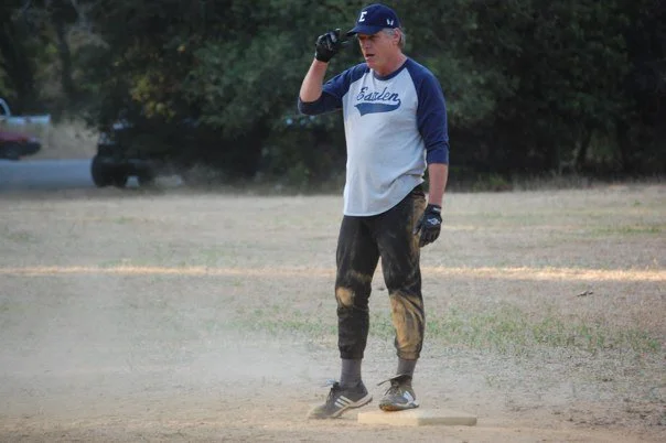 A young man in a baseball cap and long sleeves standing on a dirt field, appearing to be in a baseball game scenario.