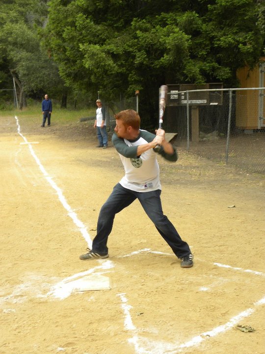 A boy swinging a baseball bat at a baseball field during a game, with two other children watching in the background and trees surrounding the field.