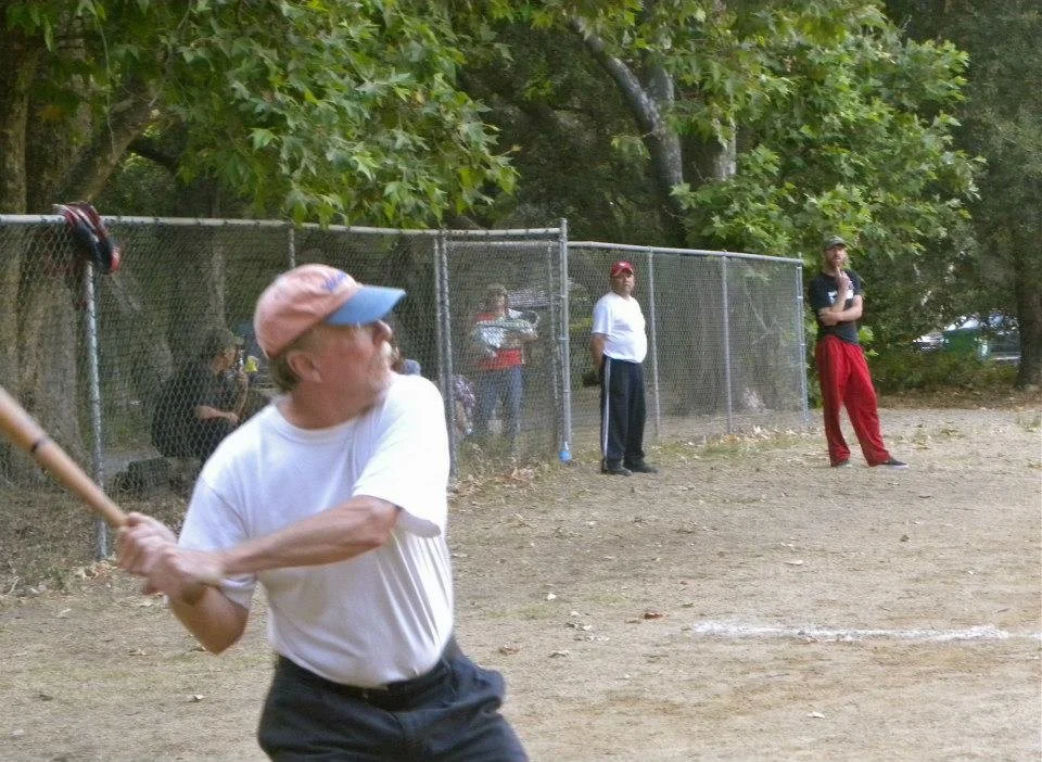 An older man in a white t-shirt and colorful cap swinging a baseball bat on a baseball field. Behind him, a chain-link fence and several people watching, some standing, some sitting, and trees in the background.