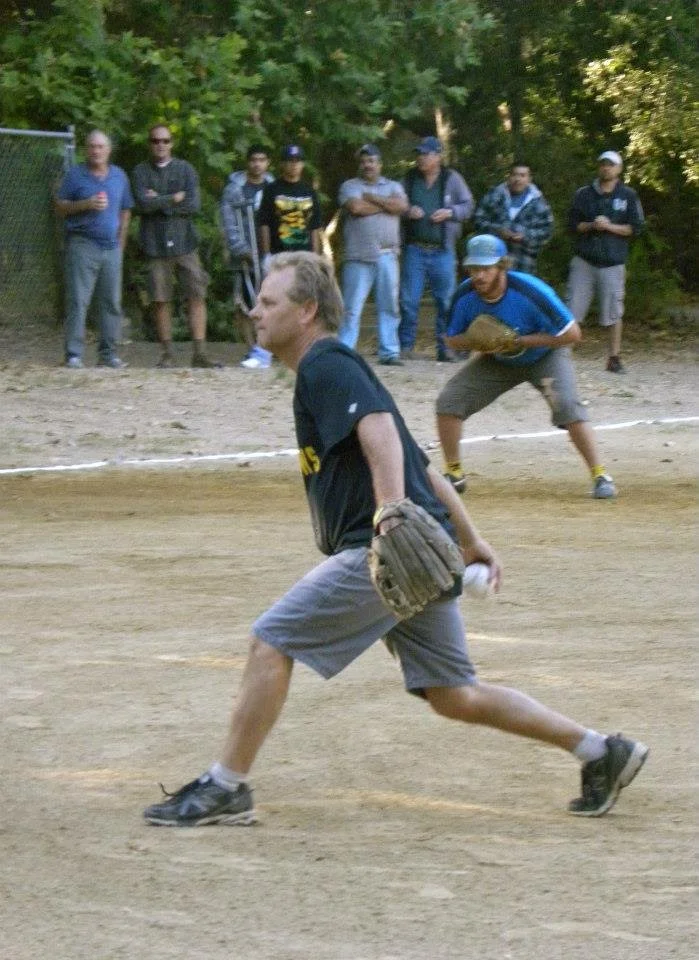 A man is fielding a baseball in a game, while another person is preparing to throw the ball, and several spectators observe from the background on a dirt field surrounded by trees.