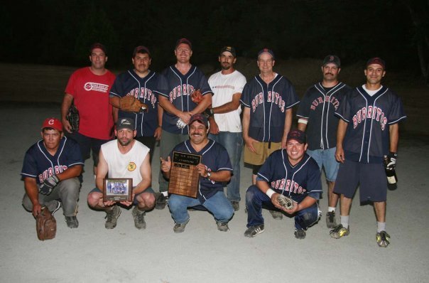 Group of nine men in baseball jerseys and casual clothing, some holding awards or plaques, on a baseball field at night.