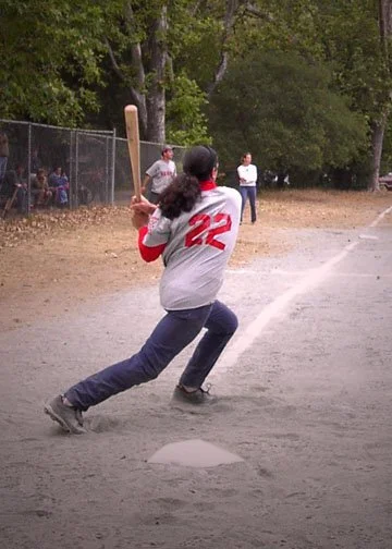A girl wearing a gray baseball jersey with the number 22 is in a batting stance on a baseball or softball field. She is holding a bat, ready to swing, with trees and spectators in the background.