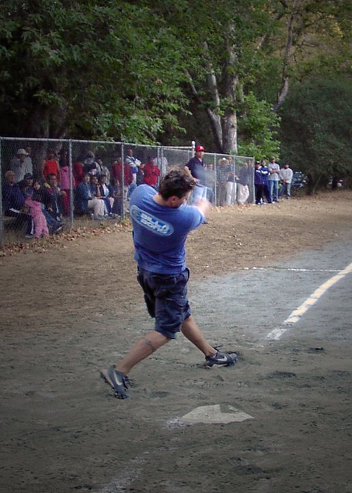 Young man swinging baseball bat at a baseball field with spectators watching behind a fence.