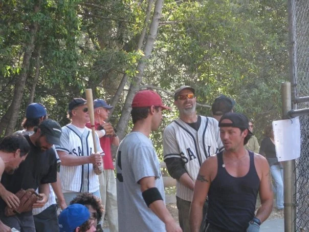 A group of men at a baseball game, some wearing baseball jerseys, gathered near a chain-link fence surrounded by trees.