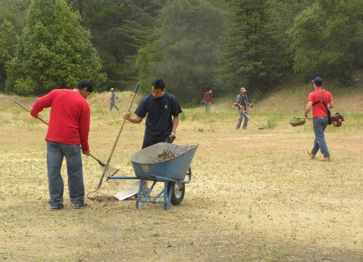 Group of people working together on a field, using shovels and power equipment, with green trees in the background.