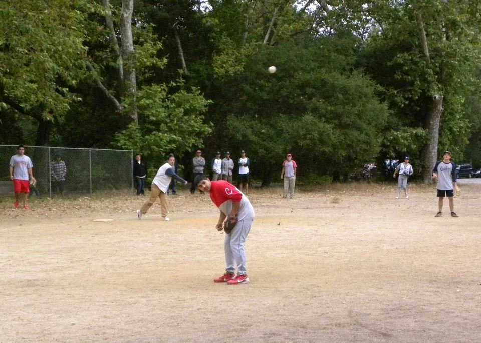 A group of people playing baseball on an outdoor field with trees in the background.