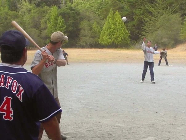 People playing baseball outdoors on a dirt field with trees in the background. One person is preparing to bat, while a person in the background appears to be pitching, and another is standing with their hand raised.