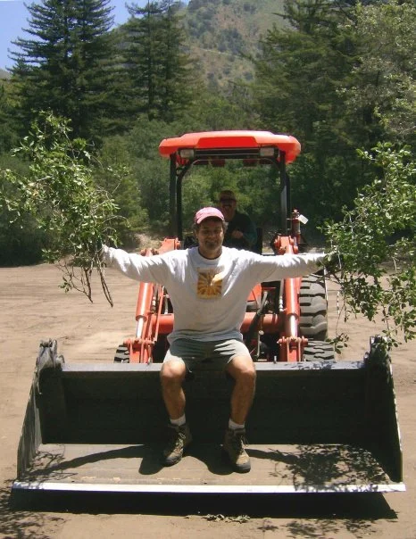 Person sitting on the edge of a front loader attachment at a construction site, with arms outstretched, in front of a tractor and surrounded by trees.