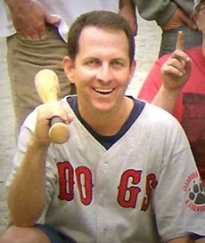 Smiling man in a baseball jersey holding a baseball bat, surrounded by people.