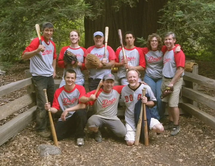Group of nine people in red and gray baseball shirts, holding baseball bats, posing outdoors on a wooded trail with trees and a wooden fence.