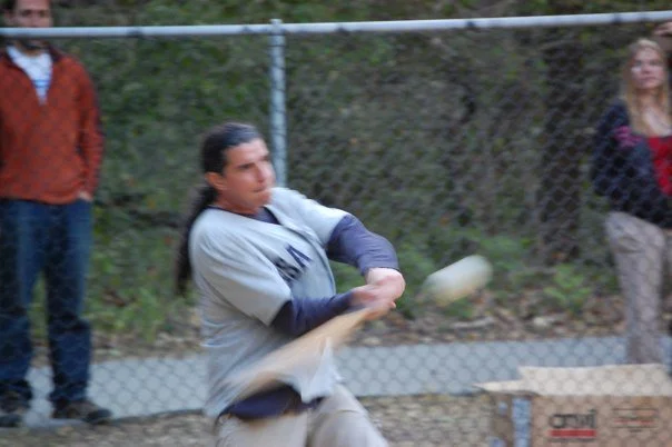 A woman in gray and navy sports uniform swings a baseball bat at a ball in a batting cage.