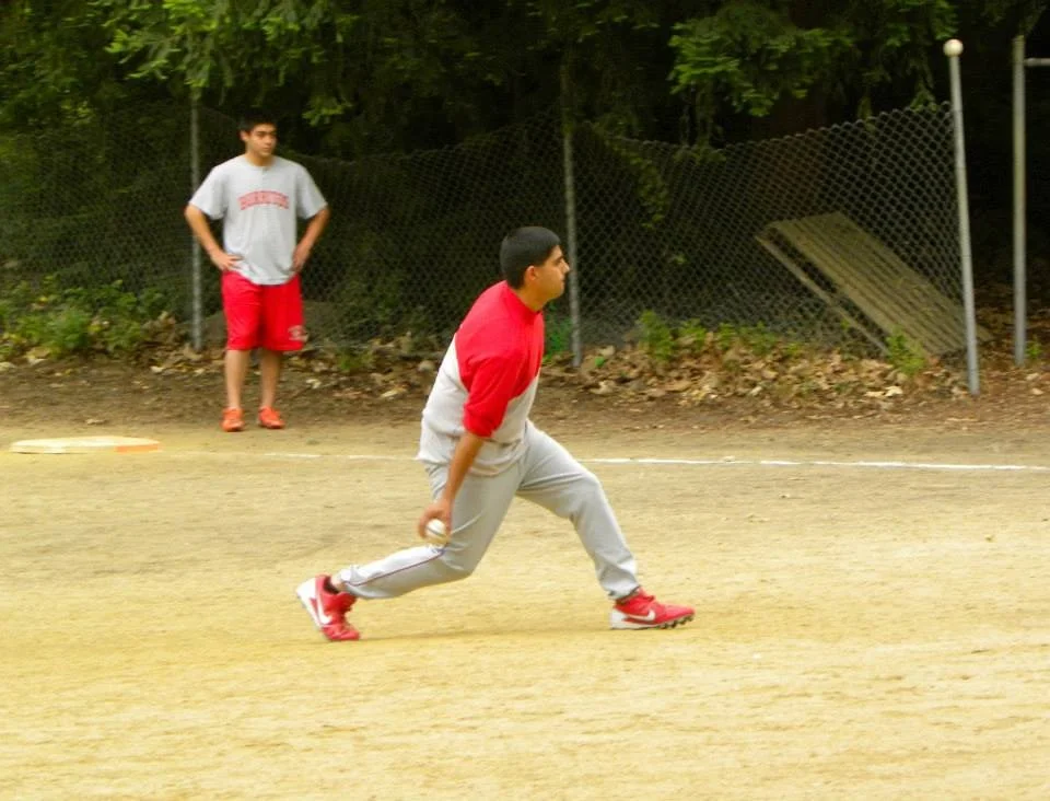 A man in a red and gray long sleeve shirt and gray pants is playing baseball, preparing to throw a ball on a dirt field. A boy in a gray shirt and red shorts is watching from behind a chain-link fence.