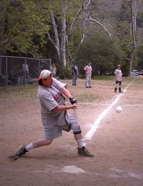 A young baseball player wearing a gray uniform, white cap, and knee brace slides into home plate at a baseball game. Several adults and children are in the background, some watching near the fence and others on the field. The scene is set in a park w