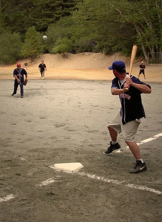 A man in a baseball uniform about to swing a bat at a ball during a game on a dirt field with trees in the background.