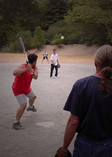 People playing baseball outdoors on a dirt field with trees in the background, a batter preparing to hit, a pitcher throwing the ball, a catcher behind the batter, and another person in the foreground.