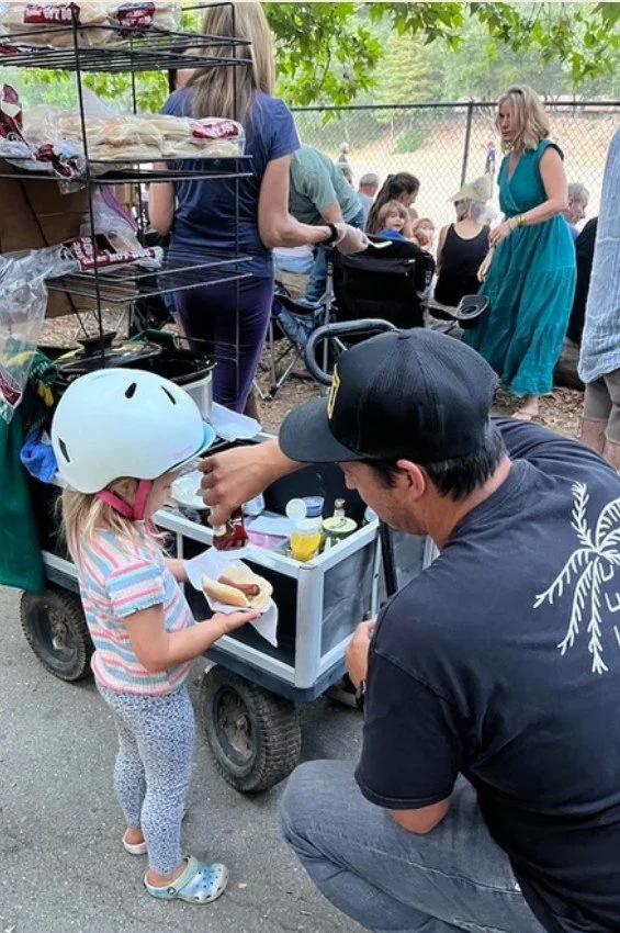 A man kneeling in front of a young girl with a bike helmet, giving her a hot dog at a community event outdoors. The girl is holding a plate with a hot dog and wearing patterned pants and a striped shirt. There are other people and a food cart in the 