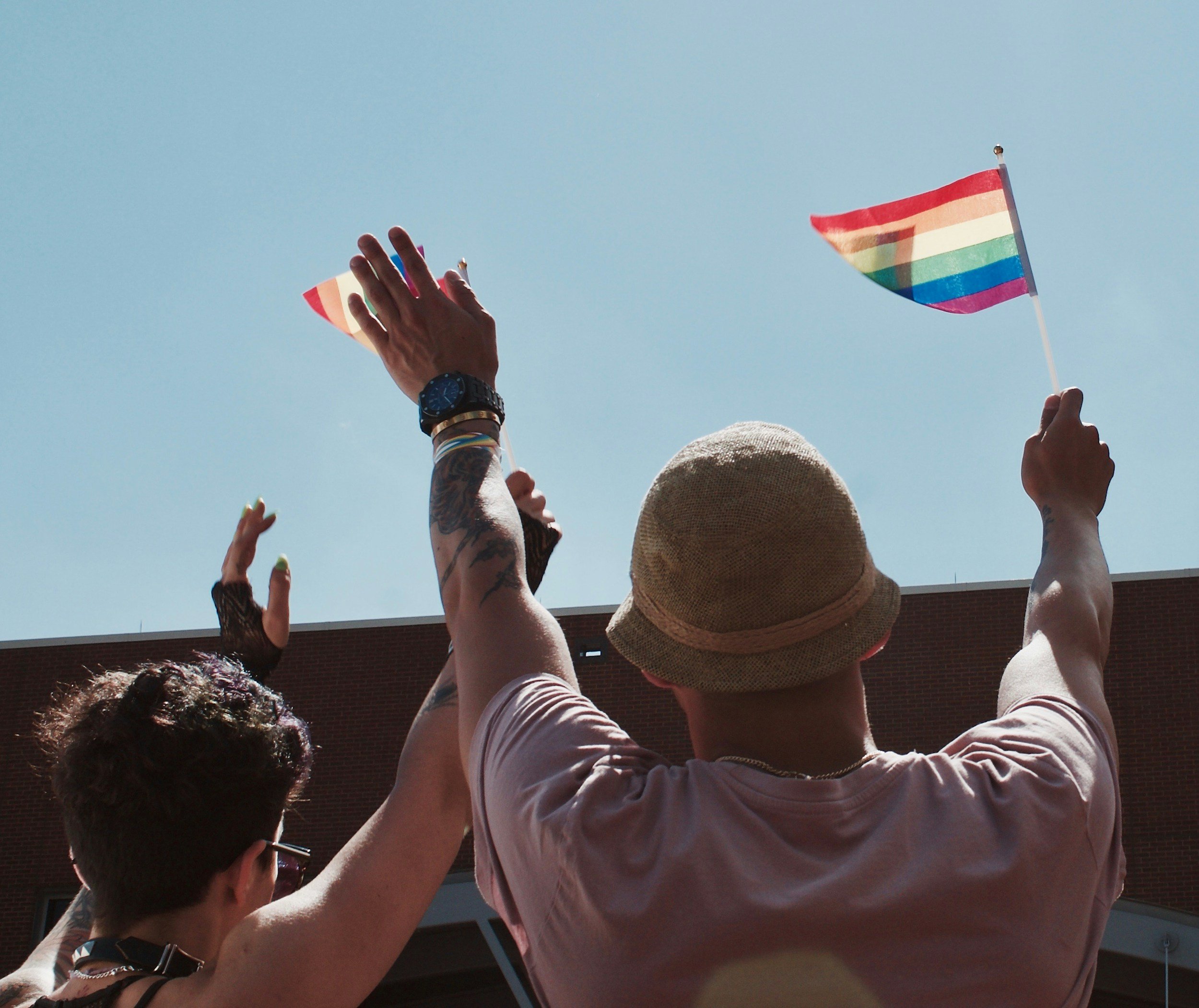 People raising hands in the air during an outdoor event, with a rainbow flag visible against a blue sky.