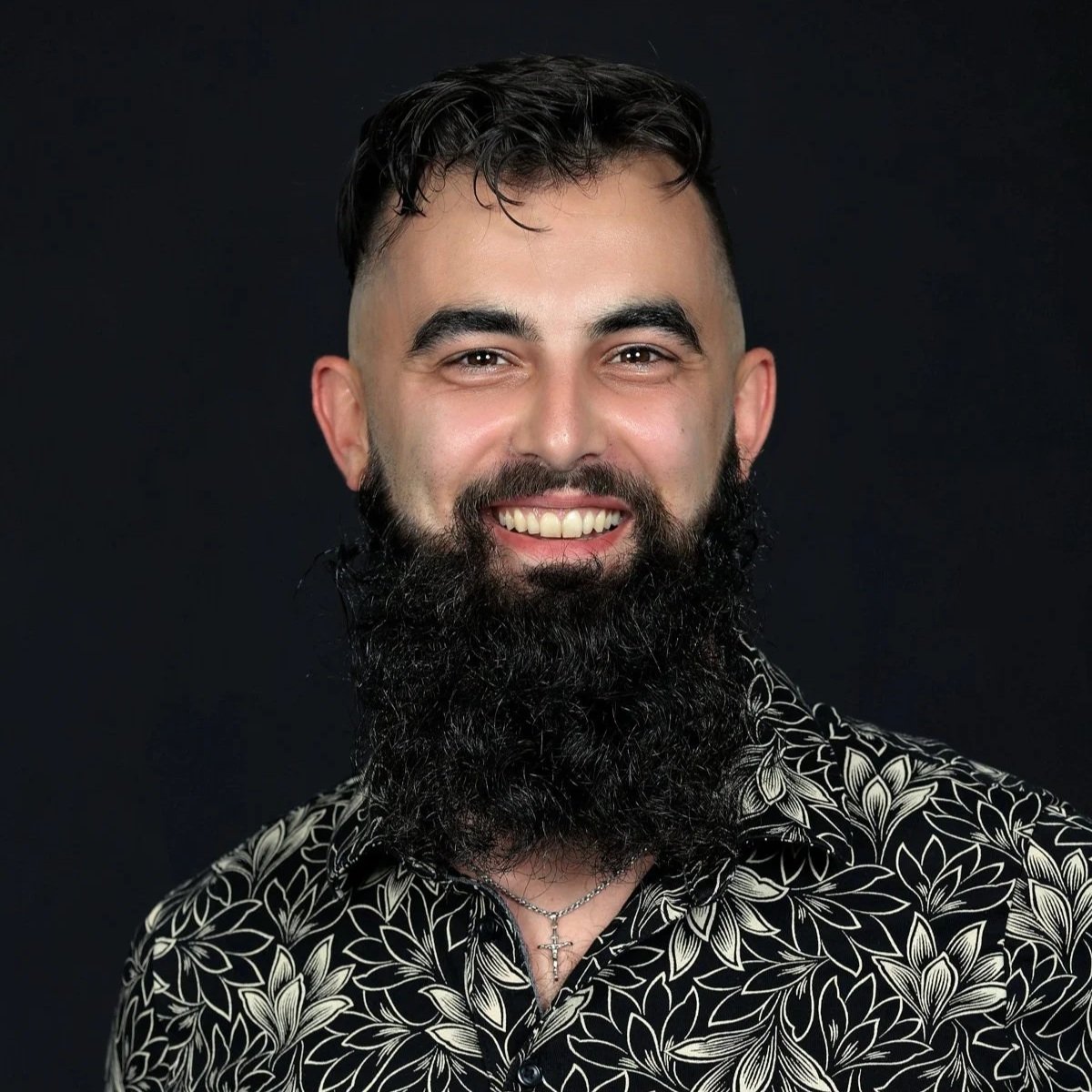 Portrait of a man with a full beard, smiling, wearing a patterned shirt and a cross necklace, with a dark background.