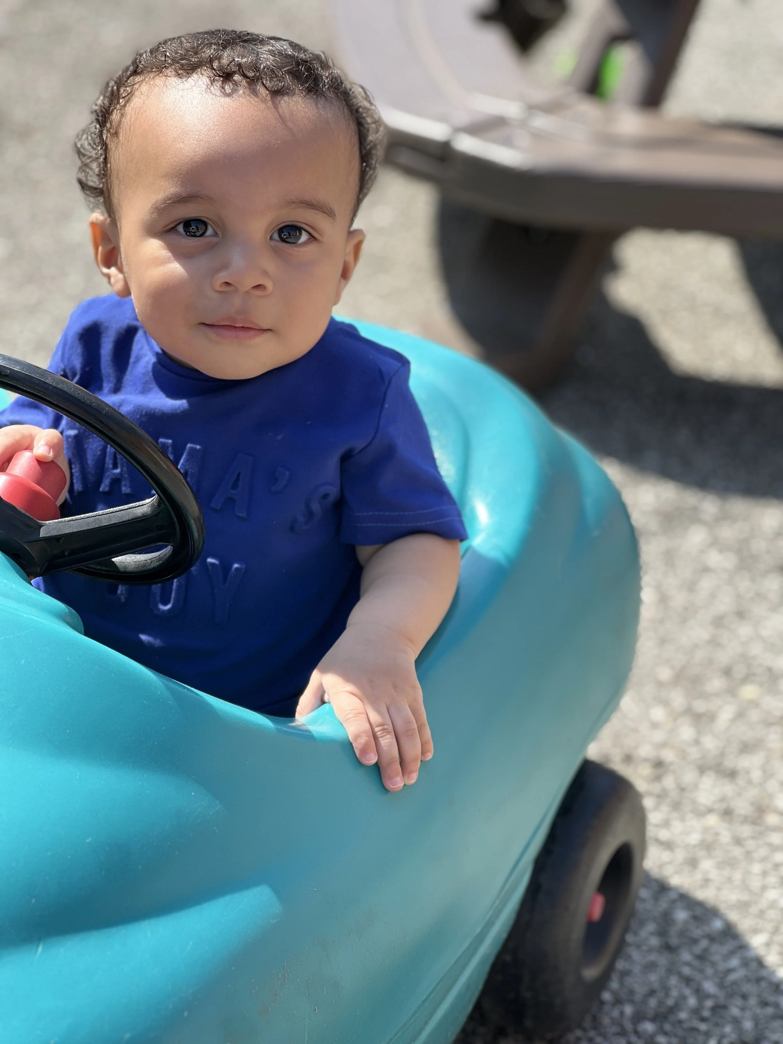 Young boy sitting in a blue toy car outdoors, looking directly at the camera with a gentle smile and large, expressive eyes.