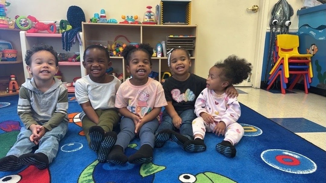 Five young children sitting on a colorful playmat in a classroom, smiling for the camera.