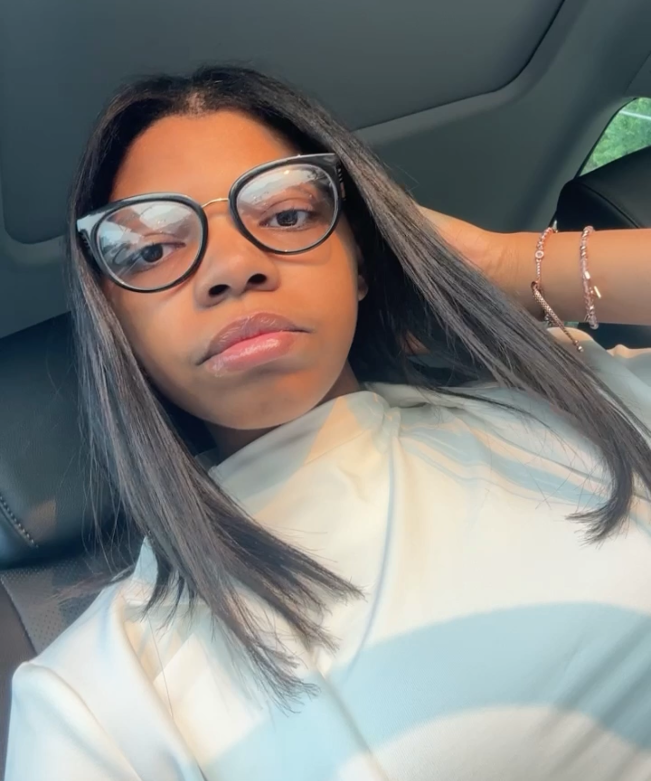 Young woman with straight hair wearing glasses and a light-colored top, sitting in a car.