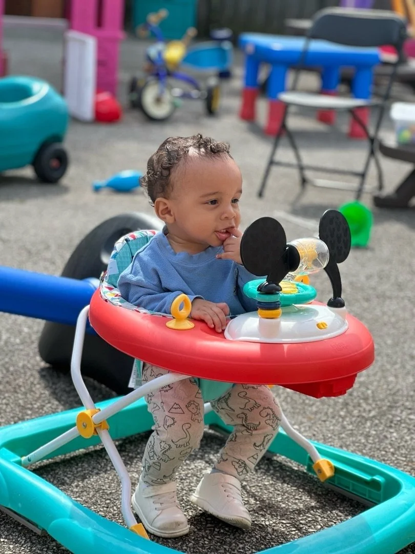 Young child sitting in a baby walker with a Mickey Mouse-themed steering wheel, outdoors in a play area with toys and colorful equipment in the background.