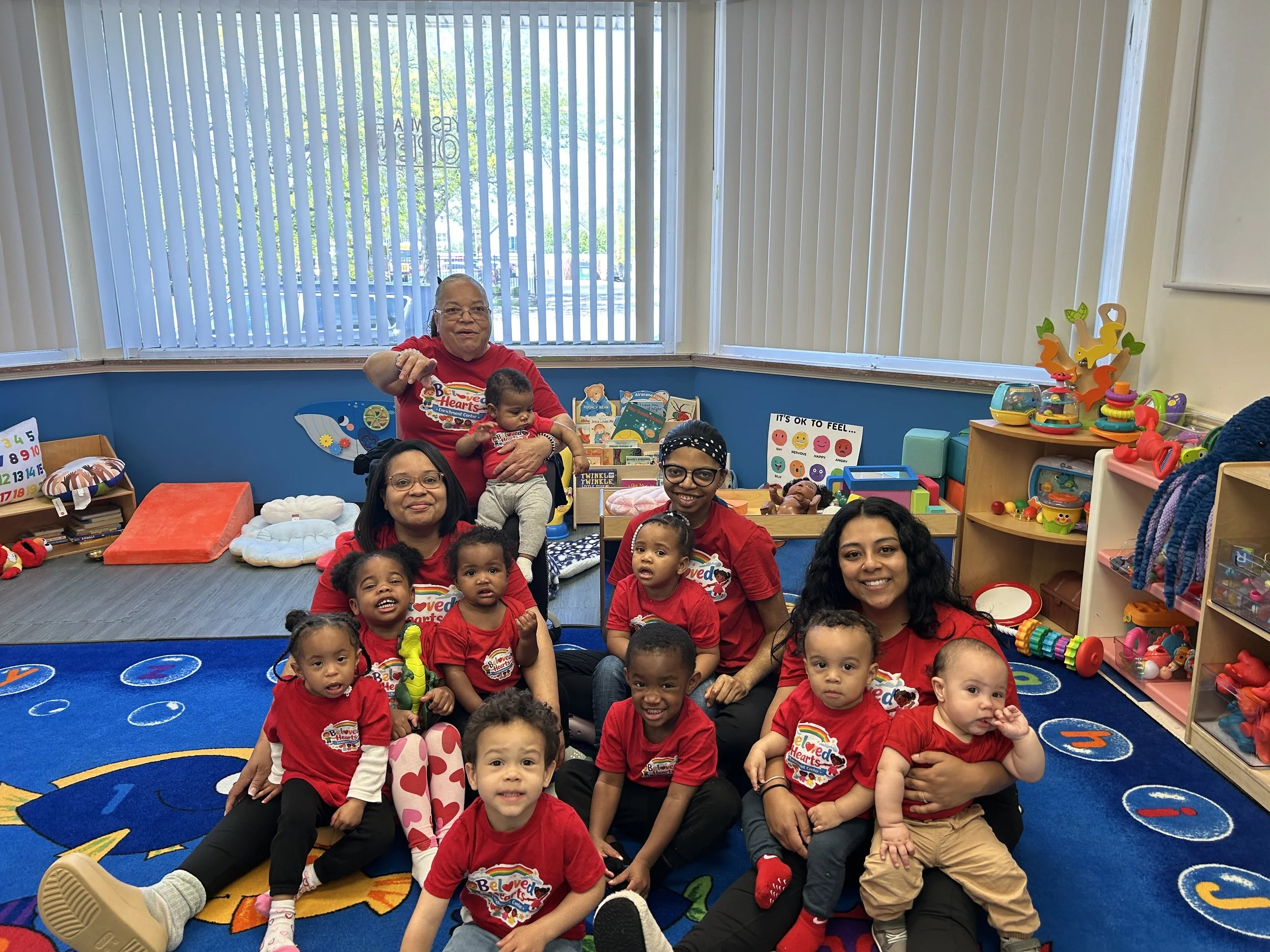 Group of children and adults gathered in a colorful indoor playroom or classroom, smiling and posing for a photo, with toys and books around them and a large window with vertical blinds in the background.