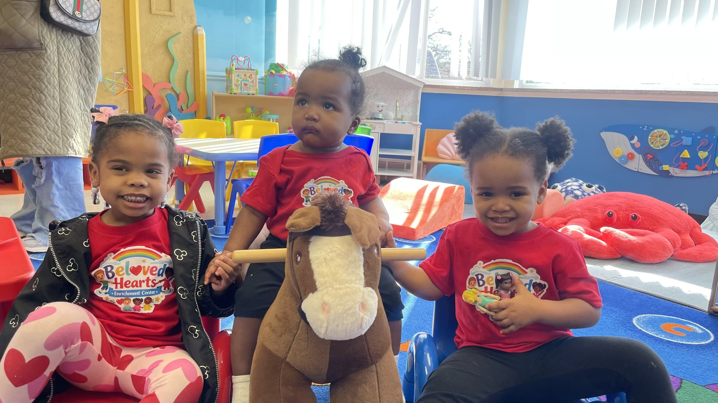 Three young children sitting at a table in a colorful indoor play area, with toys and decorations in the background. The children are wearing red shirts with 