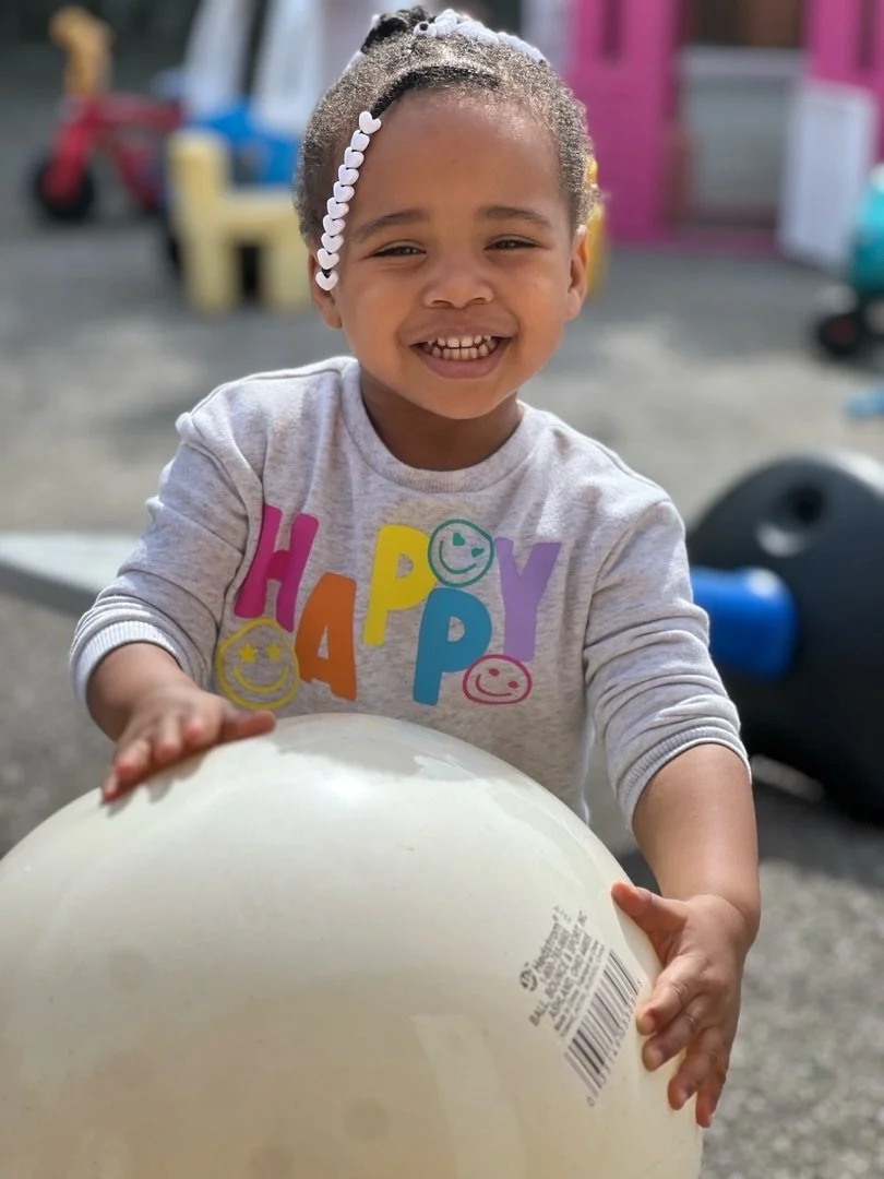 Smiling young girl playing with a white ball outdoors, wearing a gray shirt with colorful letters spelling "HAPPY" and smiling faces, with playground equipment in the background.