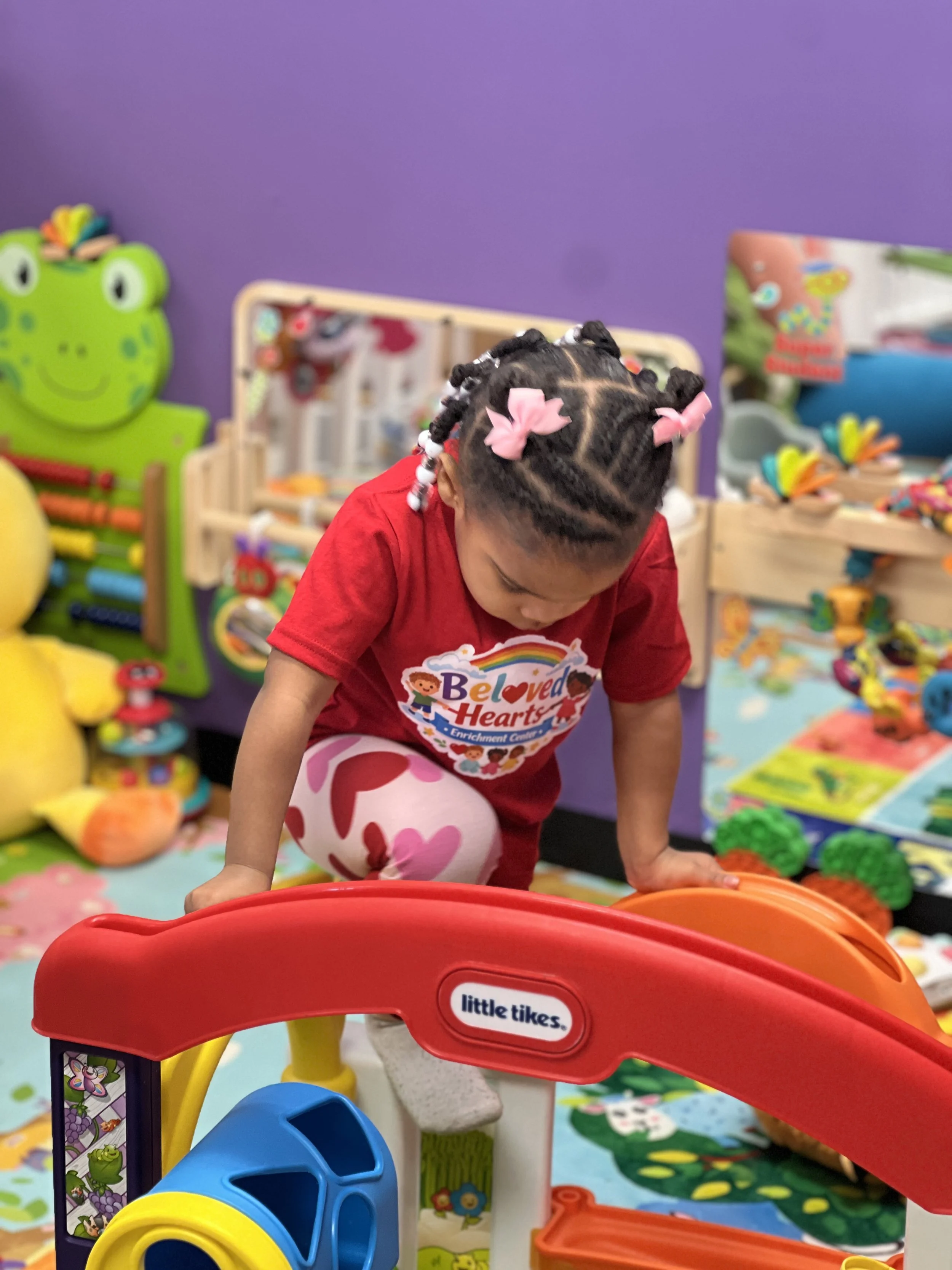 Child with braided hair wearing a red shirt and pink bows playing with colorful toy tunnel in a playroom decorated with animal-themed toys and colorful decorations.