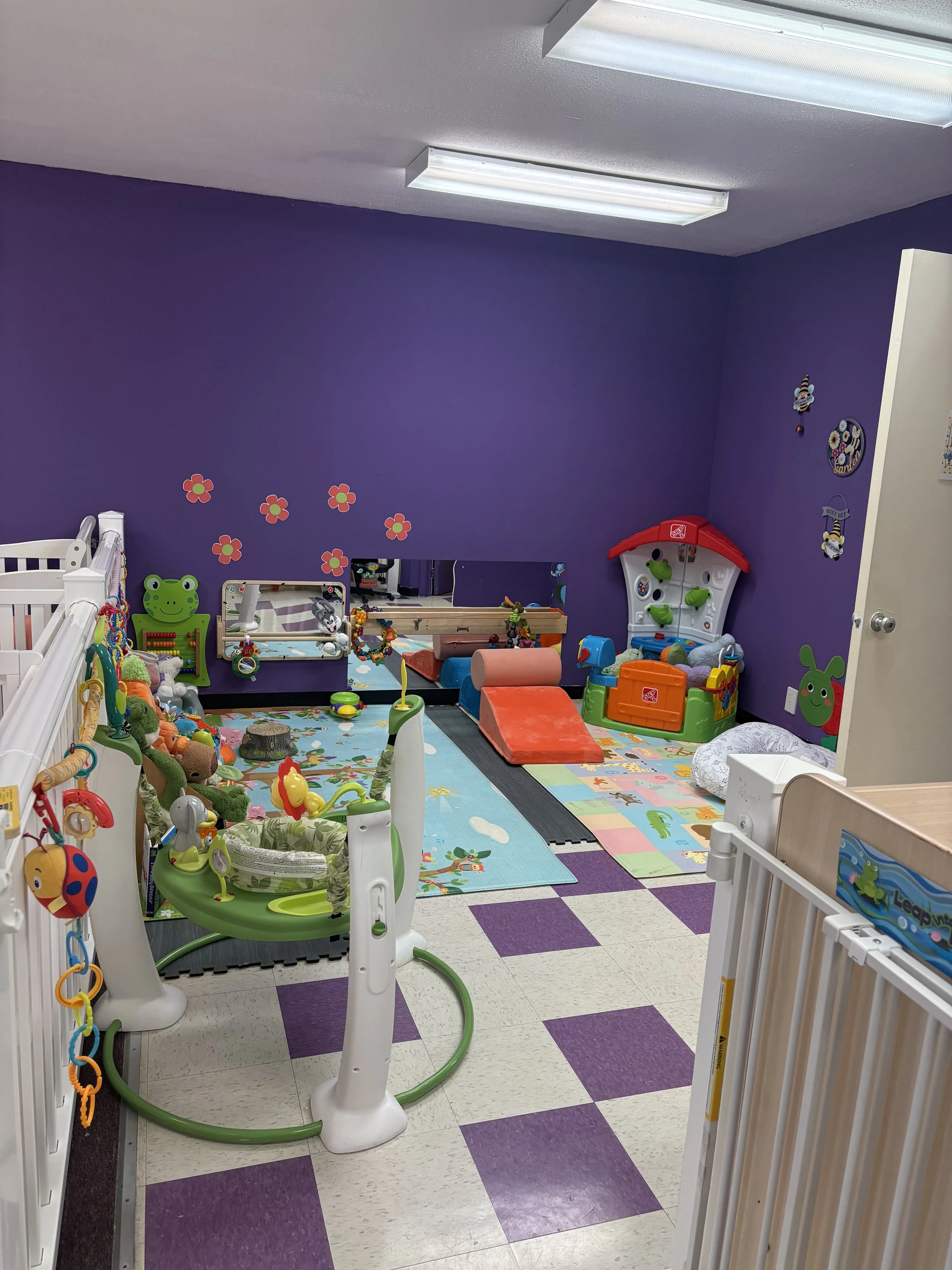 A colorful children's playroom with purple walls, decorated with flower wall stickers, featuring toys, a playmat, a baby jumper, and various stuffed animals and play equipment.