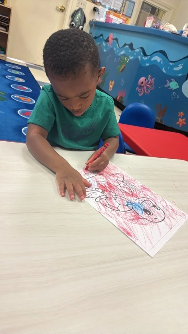 Young boy coloring with red and blue markers on a piece of paper, sitting at a white table in a colorful room with ocean-themed wall decorations.