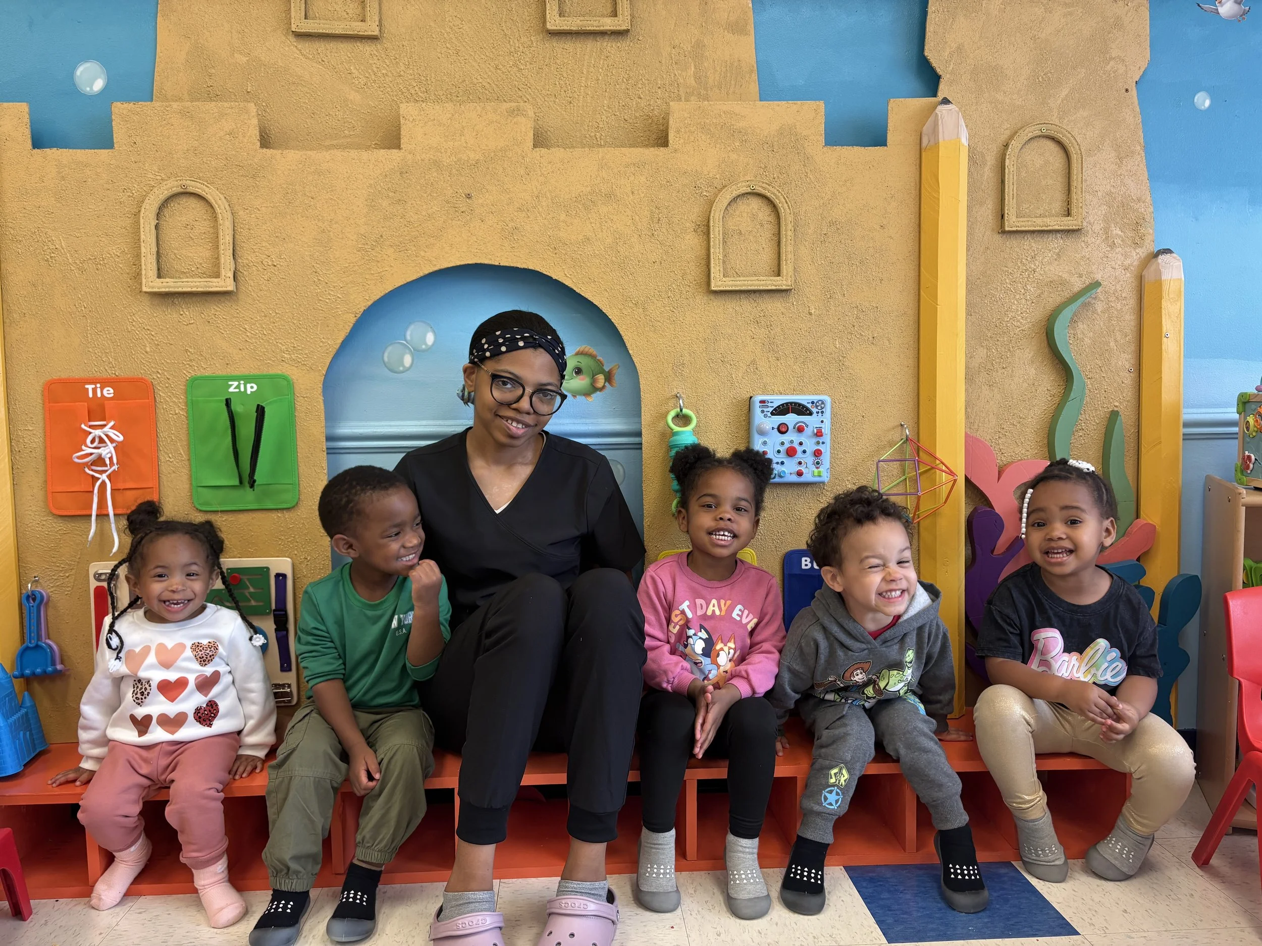 A smiling adult woman with glasses and a black headband sitting with diverse children in a colorful classroom. The background features a yellow wall with playful decorations, including window frames, a light blue painted wall, and various educational toys and art supplies.