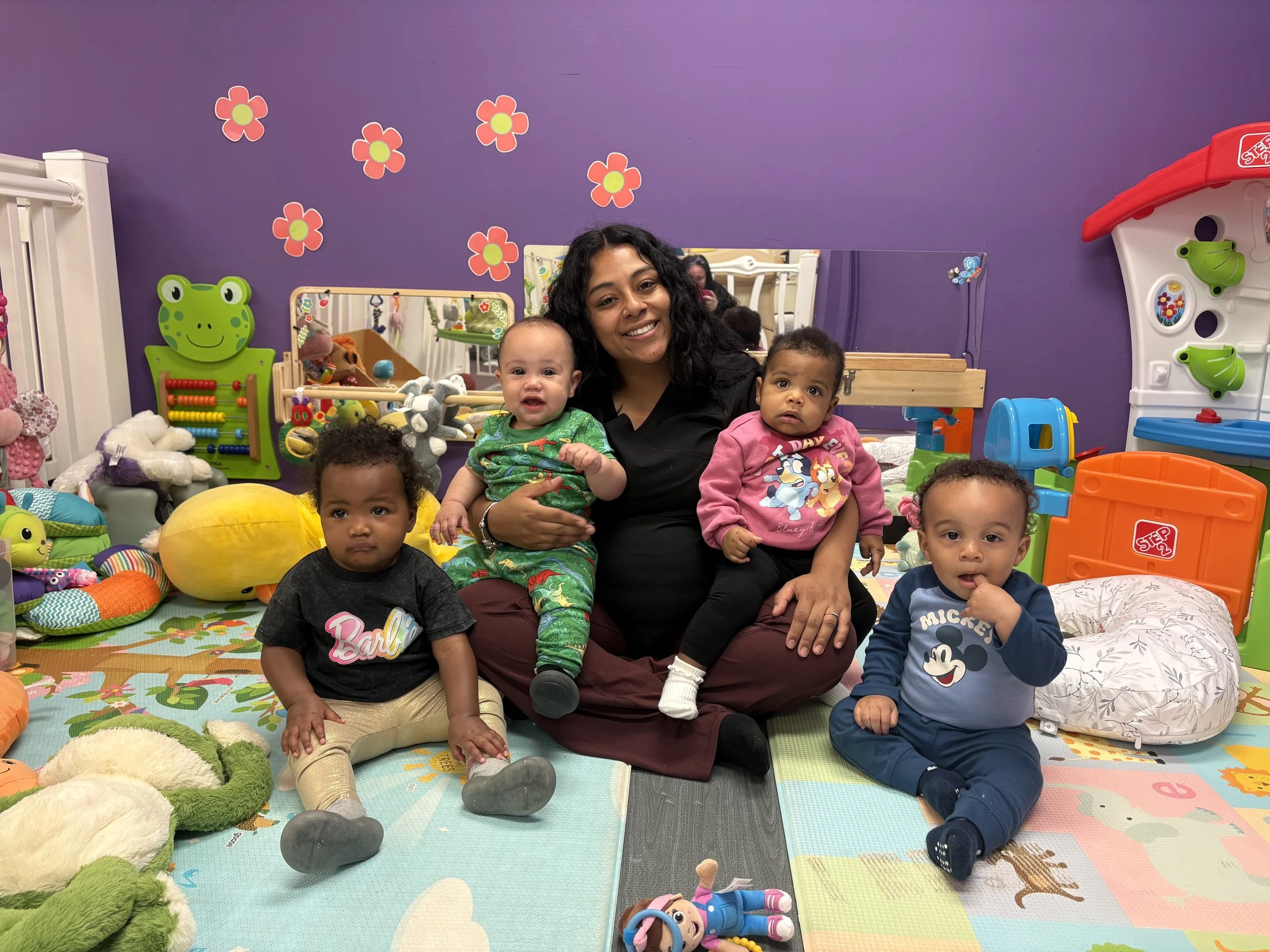A woman and five children sitting on the floor of a colorful playroom surrounded by toys and stuffed animals, with purple walls decorated with flower stickers.
