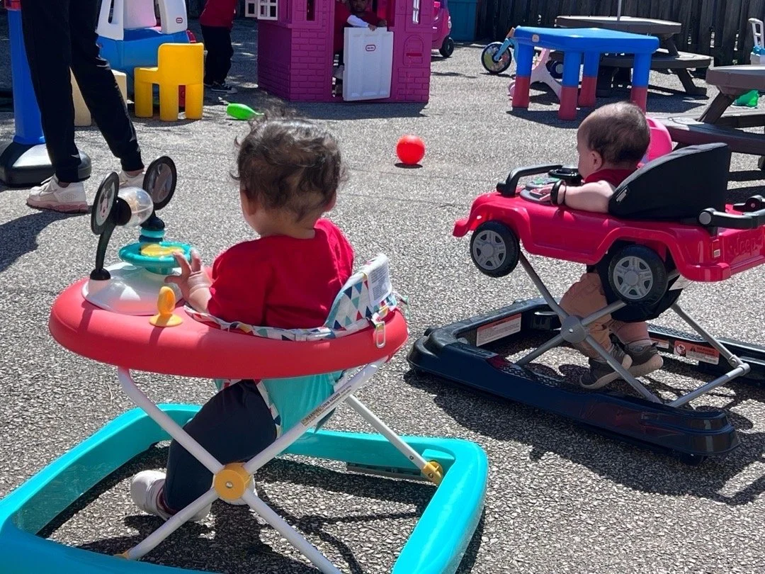 Two babies sitting in colorful baby walkers on an outdoor play area with toys and playhouses in the background.