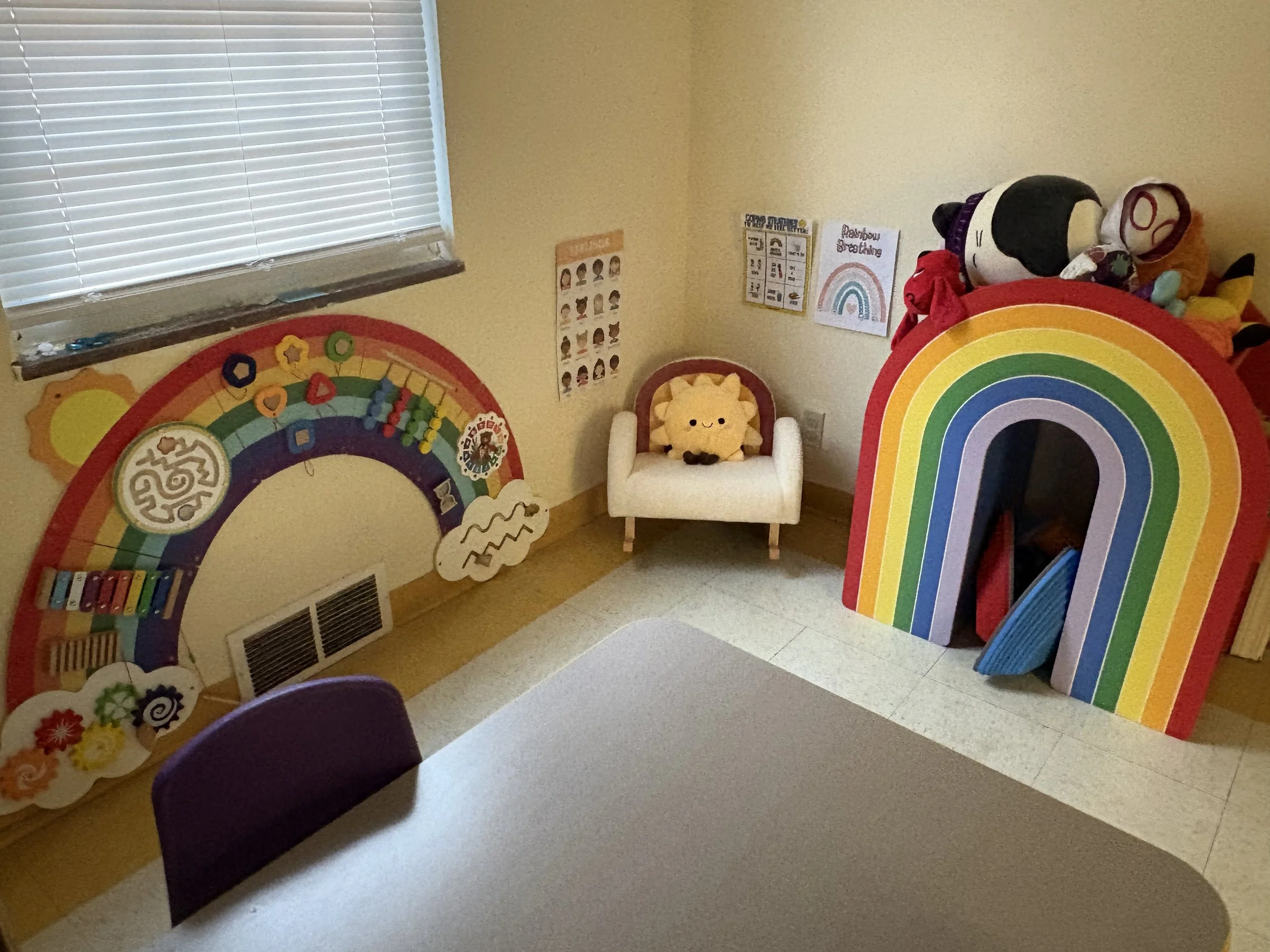 A children's playroom decorated with rainbow-themed wall art, including a large rainbow cutout and various posters, with a window with closed blinds, a small armchair, and plush toys on a rainbow-shaped shelf or play structure.