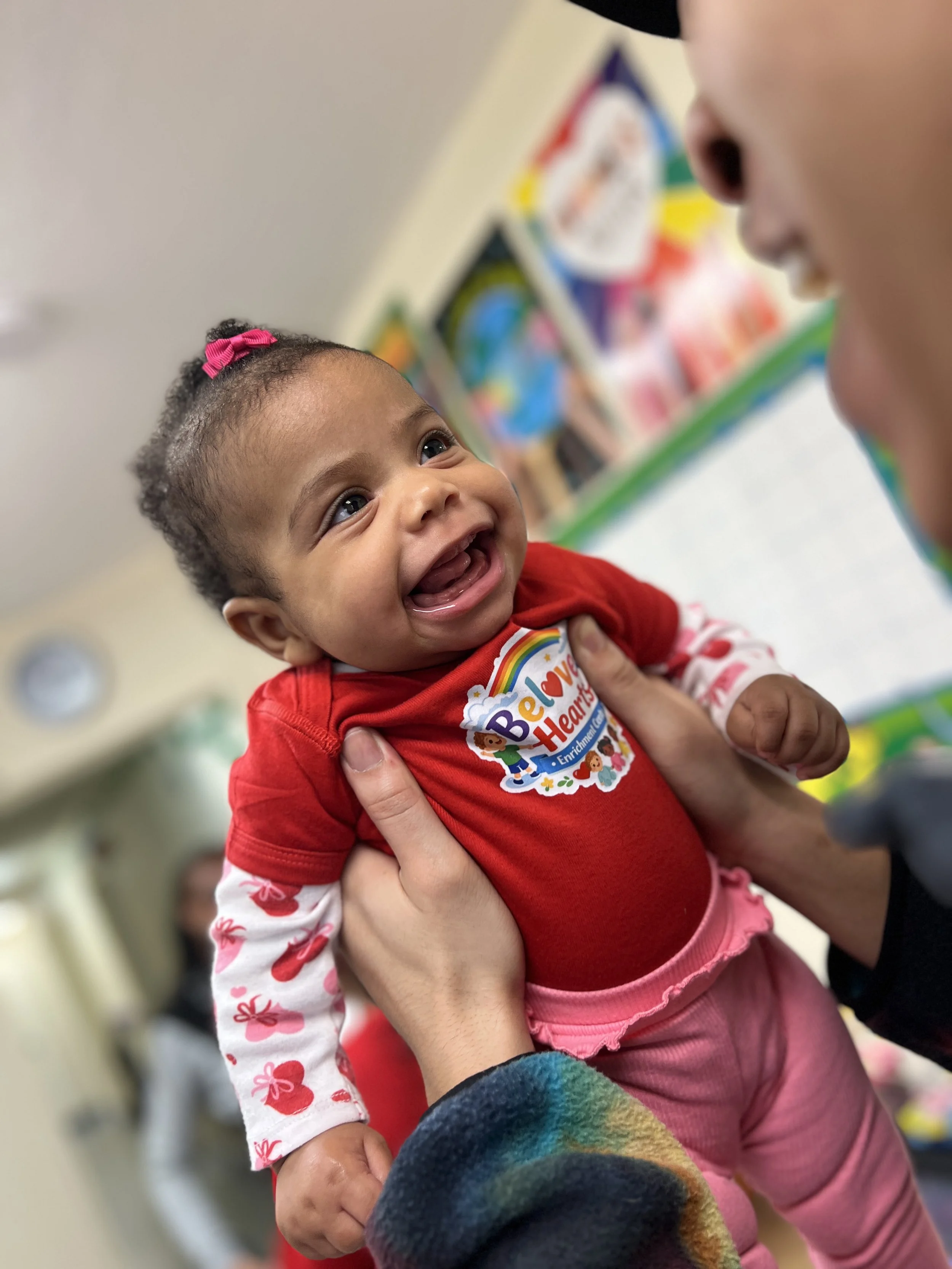 A young African American girl with a pink bow in her hair is smiling happily while being held by adult hands. The background shows colorful wall art, suggesting an indoor setting.