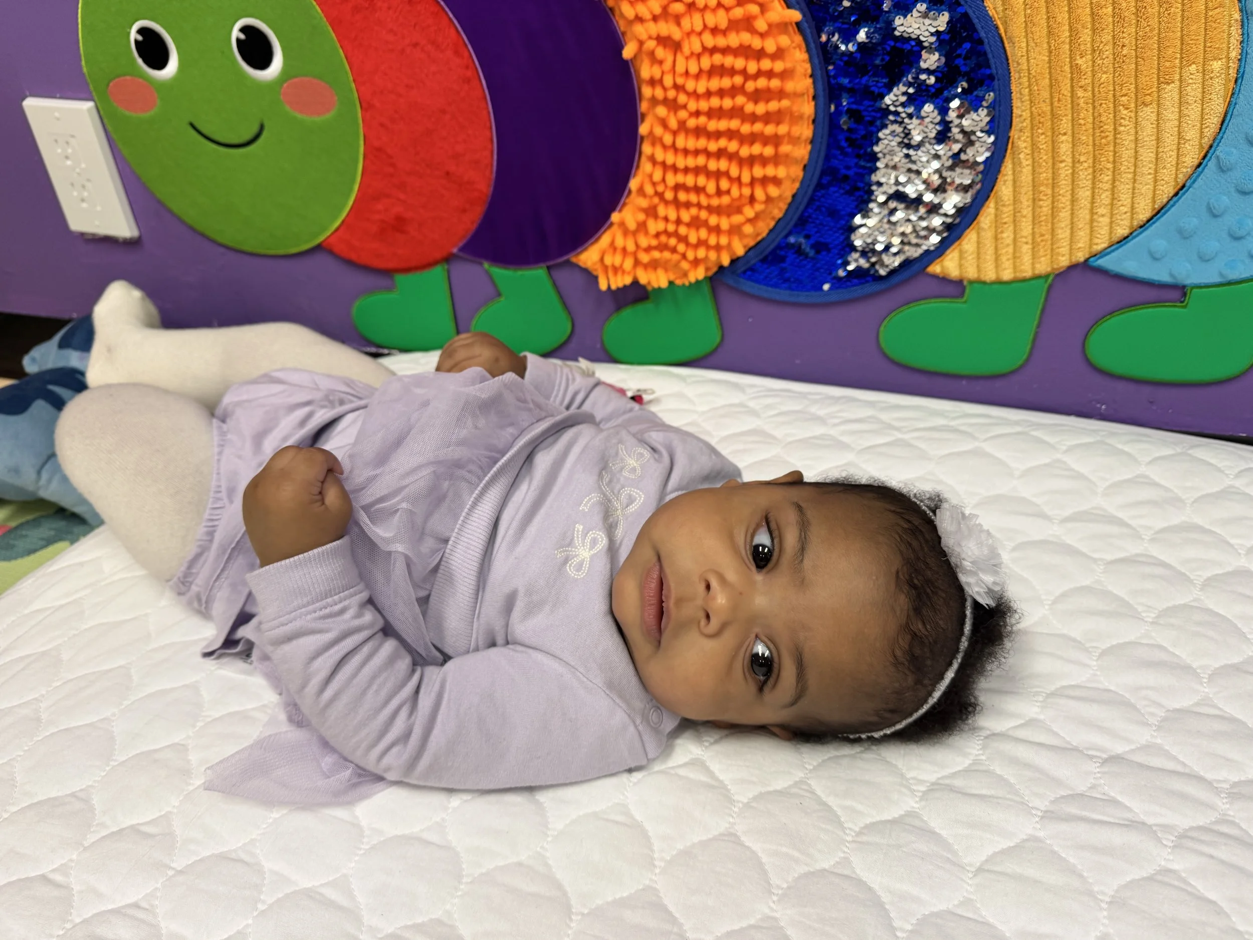 A young girl lying on a white quilted bed with a colorful wall decoration behind her. The decoration features a smiling green ladybug with large eyes, cheeks, and antennae, made of various textured and shiny materials in purple, red, orange, blue, an