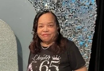 A woman smiling, wearing a black shirt with a design that includes the number 65 and a crown, standing in front of a decorative backdrop with silver and blue elements.