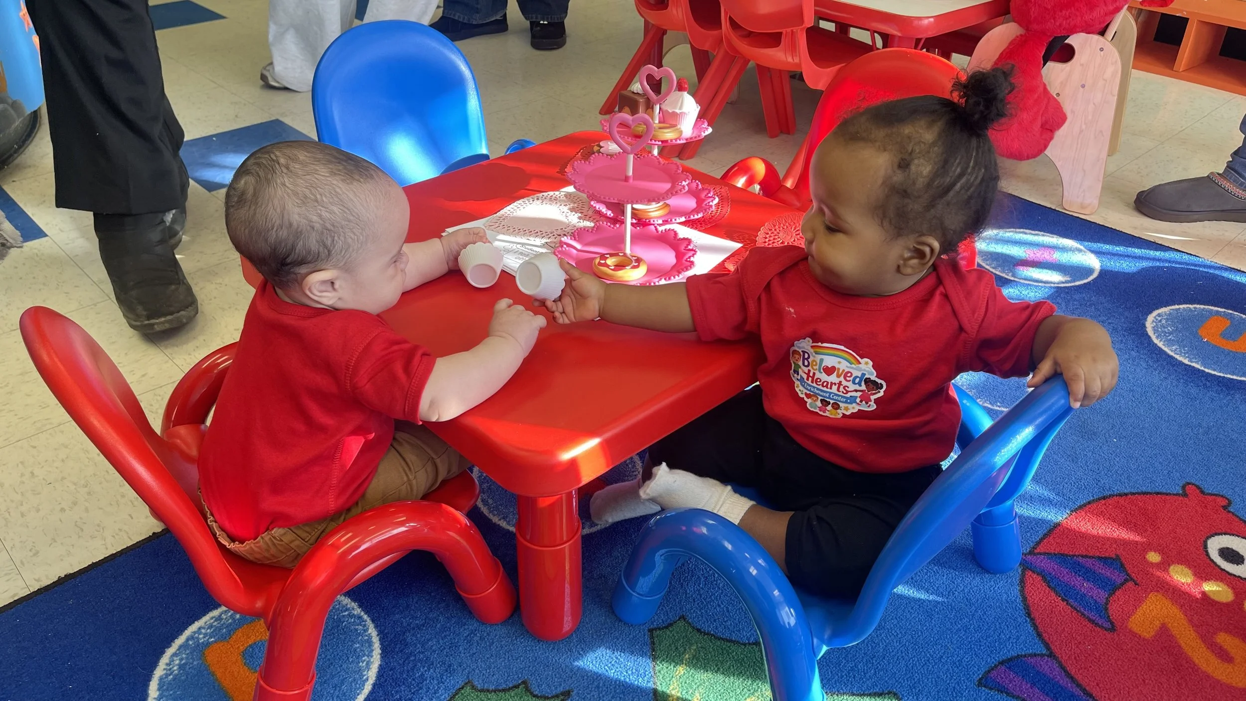 Two young children sitting at a red table, one with light skin and short hair, the other with dark skin and curly hair in a ponytail, sharing a treat. The table has pink cupcake stands with heart-shaped decorations. The children are wearing matching red shirts, and the setting appears to be a colorful indoor play area or classroom.