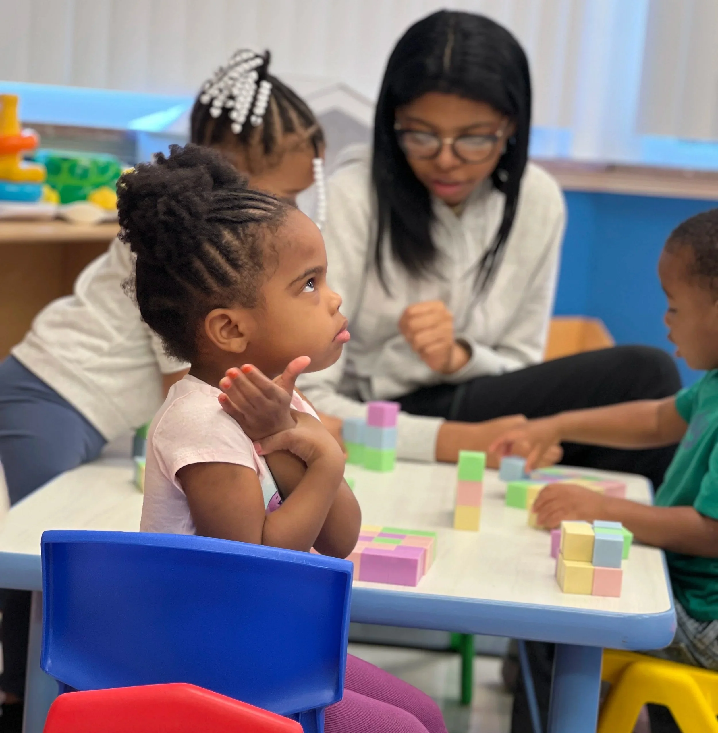 A young girl with braided hair looking up at an adult woman with glasses, sitting at a colorful table with blocks. The background shows a classroom setting.