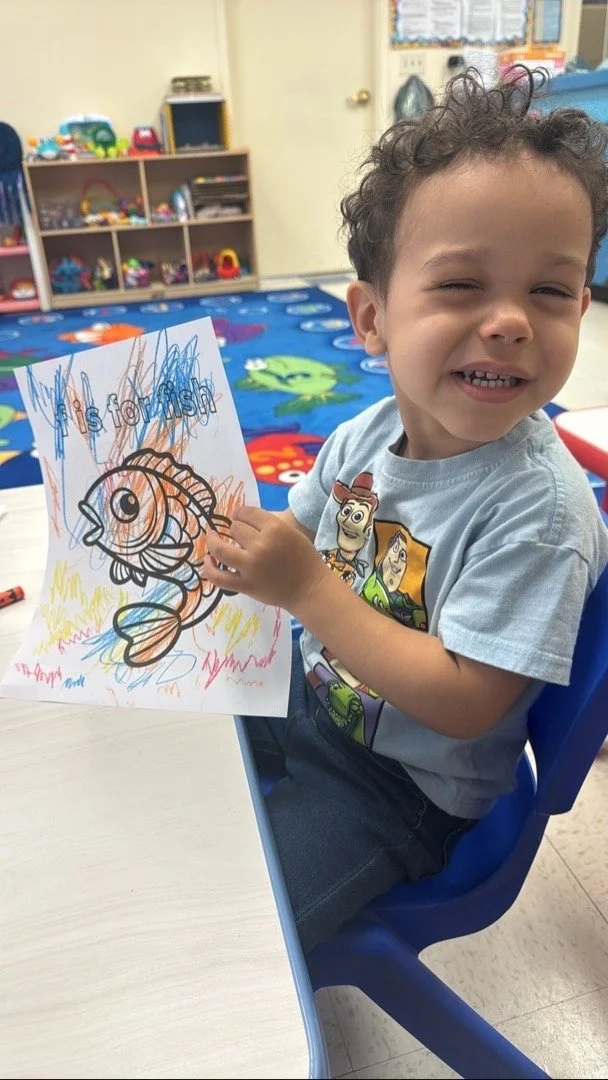 A young boy with curly hair smiling and showing a colorful drawing of a fish with the words 'Hi! Fish' written on it. He is sitting at a table in a classroom with toys and shelves in the background.