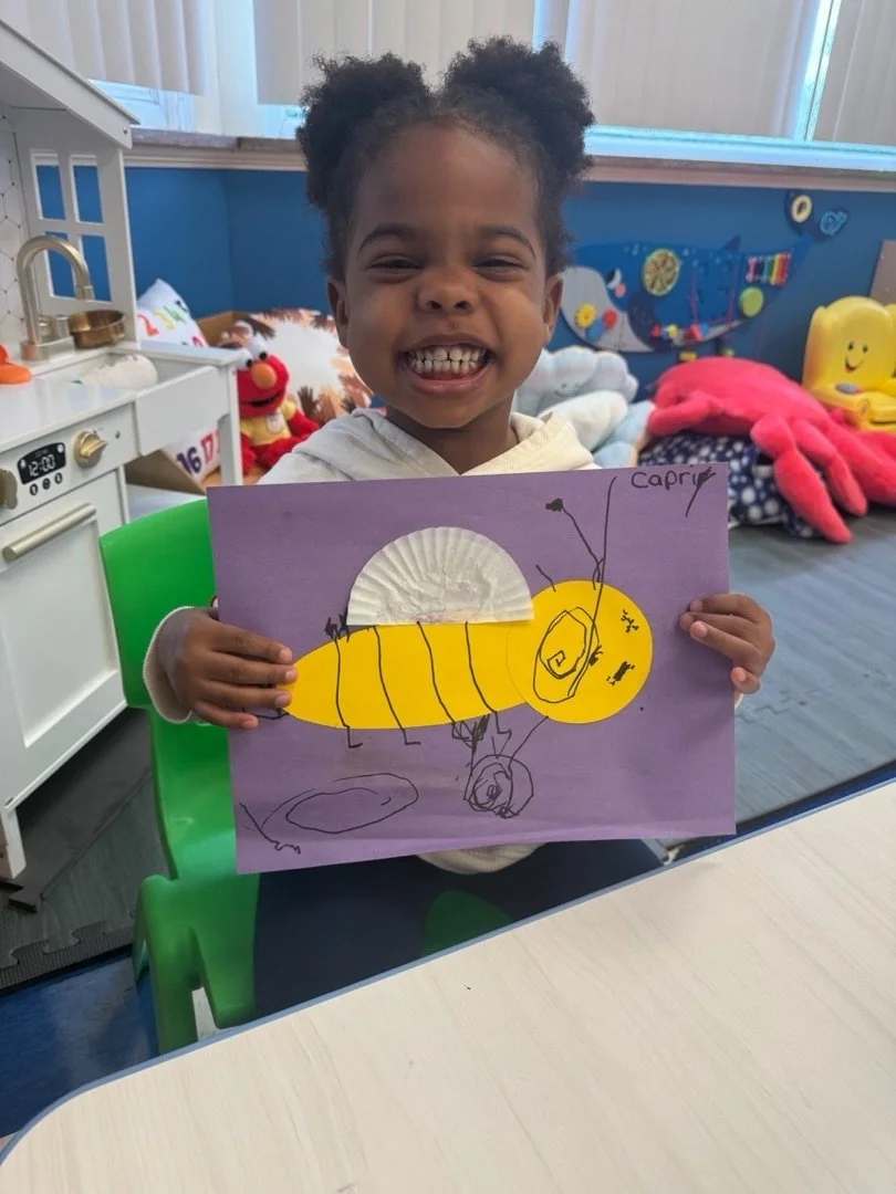 Happy young girl holding a colorful drawing of a yellow and gray caterpillar with labeled body parts, smiling widely in a playroom.