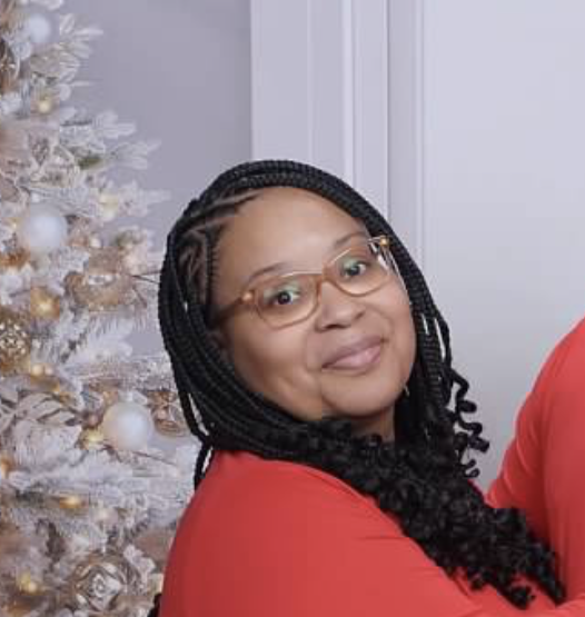 A woman with braided hair and glasses, smiling in front of a decorated white Christmas tree.